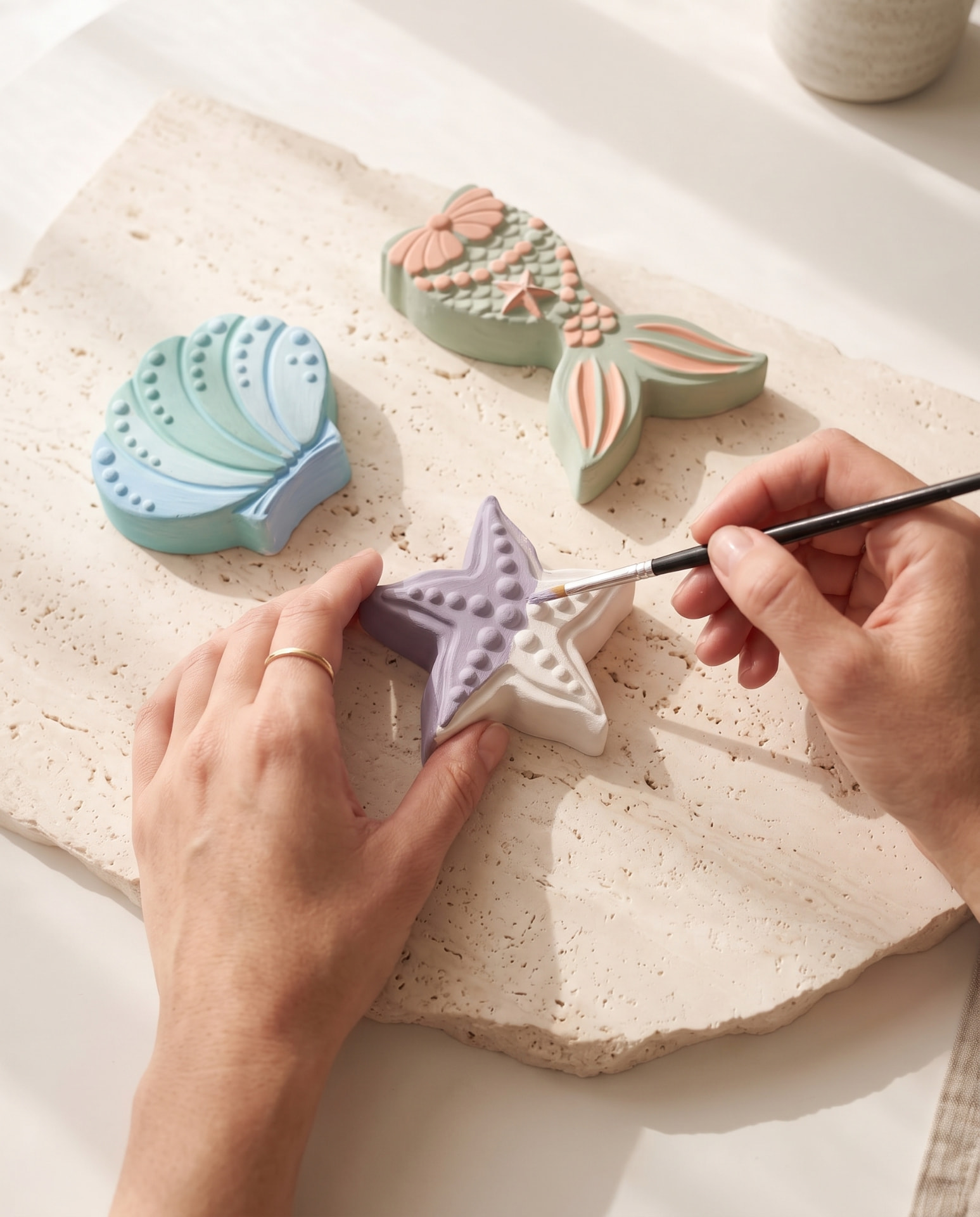 Hands painting a plaster starfish beside finished shell and mermaid-tail pieces